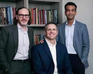 three men in suit jackets standing in front of a bookcase filled with books